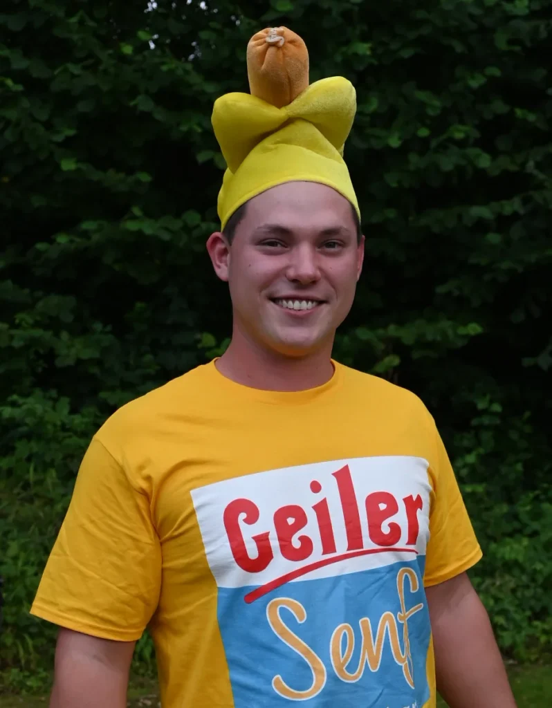 Ein junger Mann steht Portrait, sichtbar bis zu den Armen.
Er trät eine gelbe Mütze mit rießigen einem Würstchen aus Stoff.
Am Oberkörper trägt er ein gelbes T-Shirt, das einen Schriftzug mit dem Text "Geiler Senf" aufgedruckt hat.