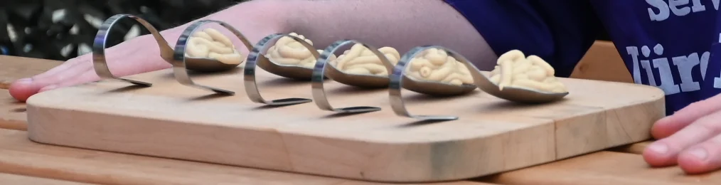 Fünf Wettkampflöffel mit je einem Häufchen Senf auf einem Holzbrett daneben je eine Hand eines Senfgladiators