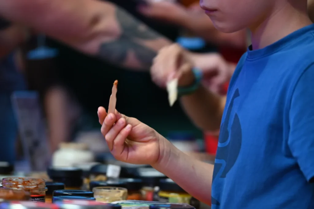 In der unteren linken Ecke einige offene Senfgläser, davor steht ein Kind, Gesicht nicht zu erkennen, man sieht den Kopf nur bis zu den Lippen. Das Kind hält einen Holzspatel in der Hand und hat damit Senf probiert.