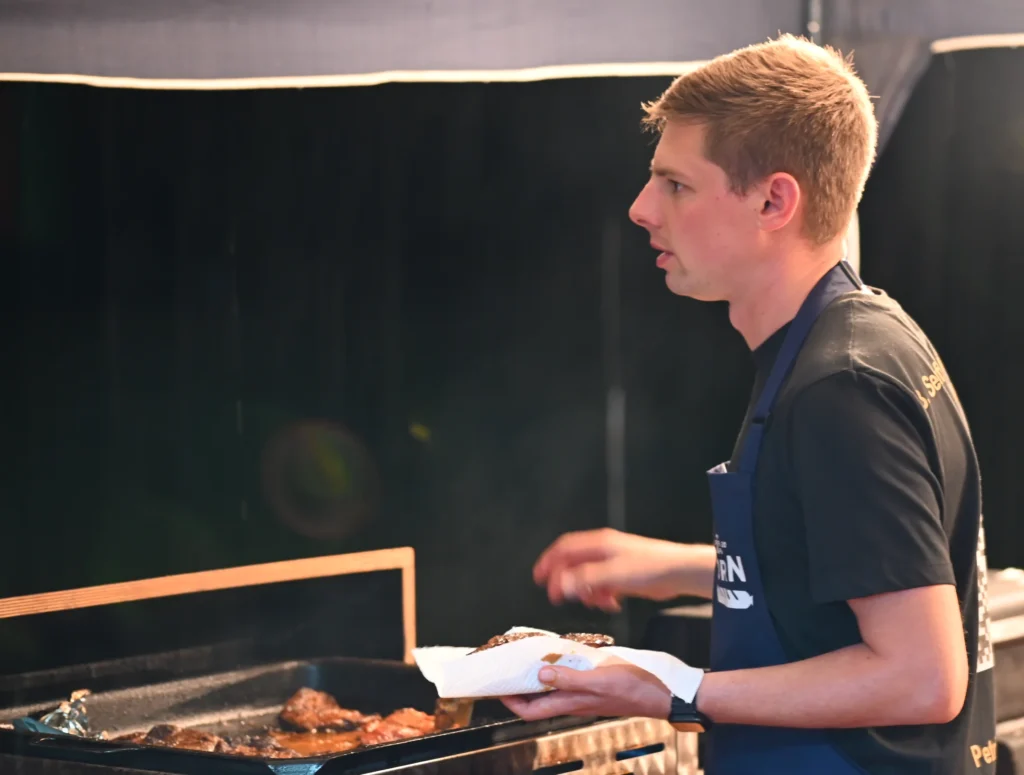 Ein junger Mann steht an einem Gasgrill mit Fleischstücken und hält ein Steak in einem Küchentuch in der Hand.
Der Mann trägt eine Schürze mit dem Aufdruck BORN.